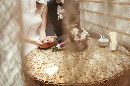 stylish bride and groom holding hands, sitting at wooden vintage table in cafe. happy wedding couple waiting for coffee, celebrationの写真素材