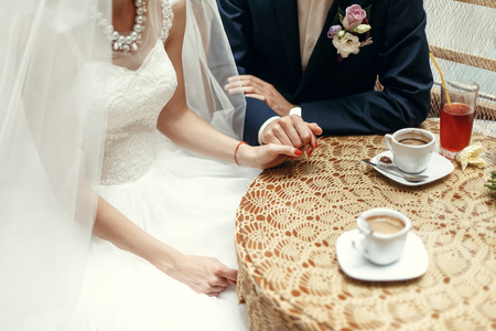 stylish wedding couple holding hands, sitting at wooden vintage table with warm coffee in cups in cafe. happy  bride and groom in dress and suit  huggingの写真素材