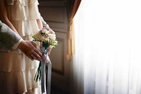 wedding bouquet. bride in vintage dress with bouquet and stylish groom embracing and hugging at window in morning light in hotel room. luxury fashionable wedding couple, sensual romantic momentの写真素材