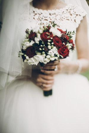 bride holding stylish red bouquet on background of her white dress in morningの写真素材