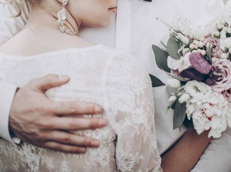 stylish wedding bride with bouquet. modern bride hugging groom, lips and earrings close up. fine art wedding photo, romantic moment, embracing newlywedsの写真素材