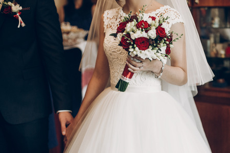 bride and groom holding stylish bouquet of red roses before wedding ceremony in church. wedding couple
