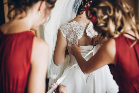 bridesmaids in red dresses helping bride put on wedding gown, morning preparation in roomの写真素材