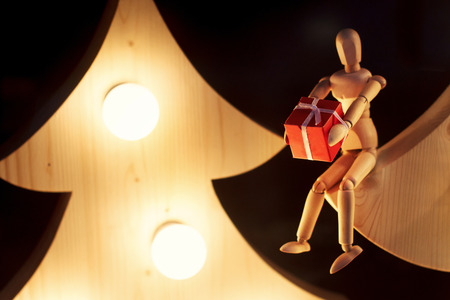 wooden toy giving gift box sitting on christmas tree with retro lights. doll of human showing present. winter holidays eco concept. space for text. greeting cardの写真素材