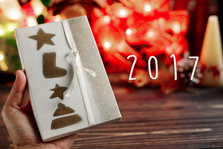2017 sign text on hand holding christmas wrapped present box on background of warm garland lights on stylish black rustic wooden background. space for text. holiday greeting card conceptの写真素材