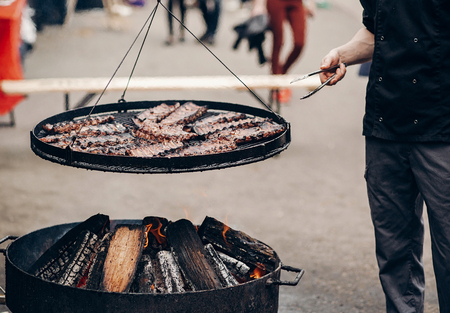 grilled meat. man holding steel tongues and roasting beef pork on big grill, under wooden fire. chef...