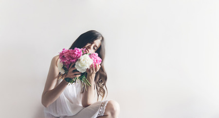 hipster woman in white dress holding pink bouquet of peonies in white room. boho bride with peony bouquet in front, relaxing in morning, space for text. sensual girl portraitの写真素材