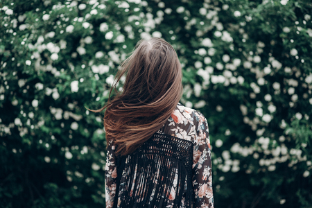 back of hipster woman posing on background of blooming bush with white flowers of spirea. boho girl waving hair in floral clothes. space for text. spring summer timeの写真素材