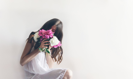 hipster woman in white dress holding pink bouquet of peonies in white room. boho bride with peony bouquet in front, relaxing in morning, space for text. spring girl with flowersの写真素材