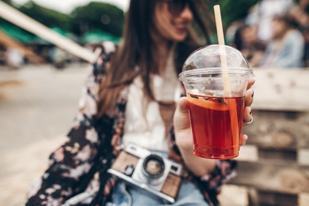 lemonade in hand. happy stylish hipster woman holding lemonade. cool boho girl in denim and bohemian clothes, with cocktail and camera at street food festival. summertime. summer vacationの写真素材