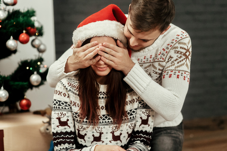 Happy family couple, handsome man preparing a christmas present surprise for beautiful woman in santa hat with closed eyes, gift card conceptの写真素材