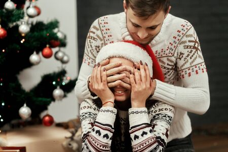 man closing eyes of his happy woman for surprising her with christmas present. cozy family moments in winter holidays at decorated christmas tree. seasonal greetings concept. space for textの写真素材