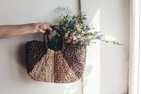 hand holding wildflowers in wicker bag at rustic window. colorful flowers in brown basket in sunlight, space for text. rural atmospheric moment. rustic wedding, creative summer pictureの写真素材