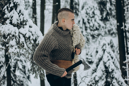 Portrait of strong viking warrior with mohawk haircut and wolf pelt armor holding axe and walking in winter woods before battle, scandinavian traditional clothing for lumberjack, viking conceptの写真素材