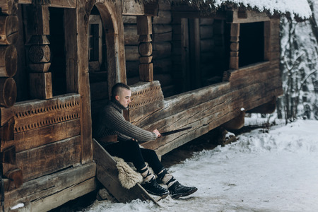 Strong viking warrior sharpening his sword while sitting near ancient wooden castle, scandinavian knight with weapon in viking costume, historical heritage conceptの写真素材