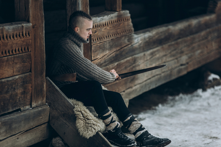 Strong viking warrior sharpening his sword while sitting near ancient wooden castle, scandinavian knight with weapon in viking costume, historical heritage conceptの写真素材