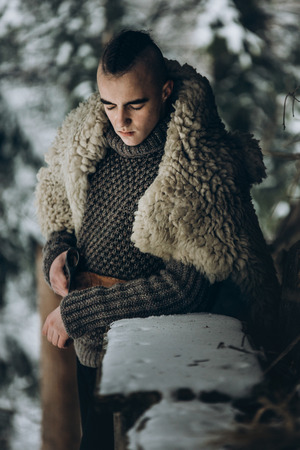 viking warrior with steel axe posing near wooden hut in scandinavian winter forest, thor viking cosplay, traditional norway costume, handsome man with mohawk hairstyle standing outdoorsの写真素材