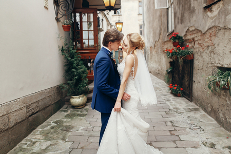 Newlywed husband and wife portrait, handsome groom in elegant blue tuxedo hugging beautiful blonde in luxury white wedding dress outdoors in old european streetの写真素材