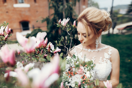 Glamourous blonde bride posing near magnolia flowers, newlywed bride portrait - beautiful woman in white dress posing near floral bush outdoorsの写真素材
