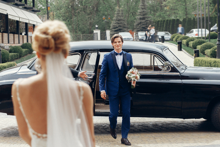Handsome happy groom with wedding bouquet walking out of black retro car and walking towards beautiful bride in wedding dress, marriage conceptの写真素材
