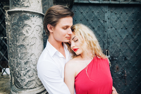 Romantic newlywed couple hugging near cathedral in Paris on honeymoon, gorgeous blonde woman in sexy red dress being embraced by handsome man in stylish shirt, family portrait conceptの写真素材