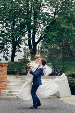 Happy newlywed couple portrait, strong handsome groom lifting up bride in the air, groom carrying beautiful bride outdoors, car in the backgroundの写真素材