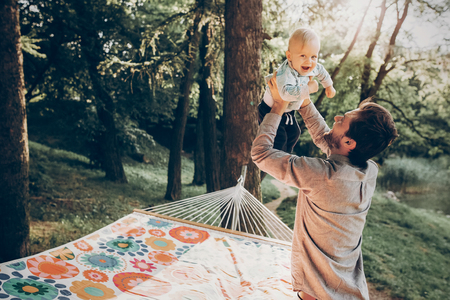 Happy hipster father playing with his son in a park near a hammock while on camping trip in the woods, family concept, handsome man holding baby outdoorsの写真素材