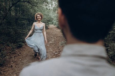stylish hipster bride and groom walking and dancing in green summer forest. happy couple in love, modern outfit, relaxing at park. girl in dress and straw hat with peony. rustic wedding conceptの写真素材