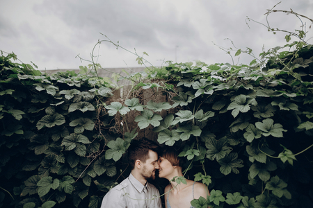stylish hipster bride and groom kissing in green leaves, holding hands. happy couple embracing, in love relaxing in summer park, picnic date. girl in fashionable modern dress. rustic weddingの写真素材