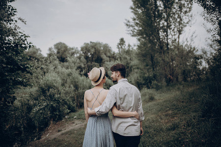 stylish hipster couple walking and hugging, back view. man and woman in modern outfit, in love relaxing in summer park. girl in dress and straw hat with peony. rustic wedding conceptの写真素材