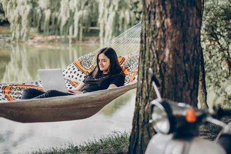 Young female freelancer working on laptop while lying on a hammock near a lake, hipster woman resting in nature near retro italian motorcycleの写真素材