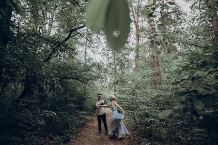stylish hipster couple walking and dancing in green summer forest. happy man and woman in love, modern outfit, relaxing at park. girl in dress and straw hat with peony. rustic wedding conceptの写真素材
