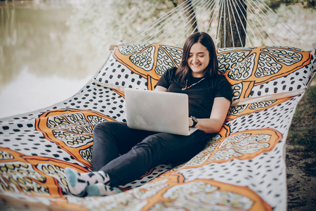 Beautiful brunette woman relaxing on hammock outdoors near lake in the forest, freelancer working in the park while resting in hammock, freelance conceptの写真素材