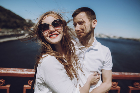 happy woman with windy hair in sunglasses smiling, stylish couple in love having fun on bridge in the summer city. modern woman and man in fashionable white clothes embracing at the riverの写真素材