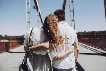 stylish couple in love hugging, back view with windy hair, on bridge in the summer city. modern woman and man in fashionable white clothes embracing at the river, romantic momentの写真素材