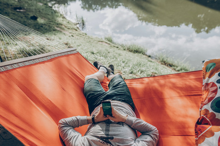 hipster man holding smart phone with empty screen with space for text, and relaxing in hammock in sunny summer park. mock up. guy looking at blank phone, technology communication conceptの写真素材