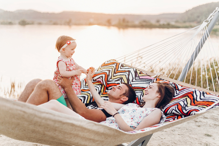 happy stylish family with cute daughter relaxing in hammock on summer vacation in evening sun light on the beach. hipster couple with child resting and having fun in sunsetの写真素材