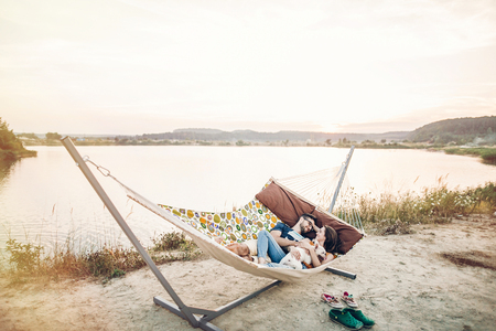 happy hipster couple with bulldog relaxing in hammock on the beach in sunset light, summer vacation. stylish family with dog cuddling and having fun, cute moments in summer eveningの写真素材