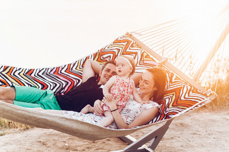 happy stylish family playing with cute daughter, relaxing in hammock on summer vacation in evening sun light on the beach. hipster couple with child resting in sunset. space for text. summer holidayの写真素材