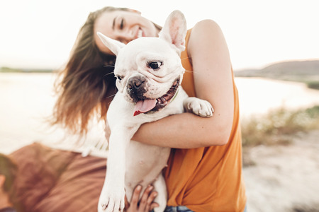 happy hipster woman playing with bulldog on the beach in sunset light, summer vacation. stylish girl with funny dog resting, hugging and having fun in sun, cute moments. space for textの写真素材