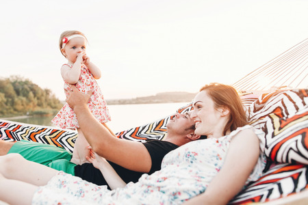 happy stylish family playing with cute daughter, relaxing in hammock on summer vacation in evening sun light on the beach. hipster couple with child resting and having fun in sunset. space for textの写真素材