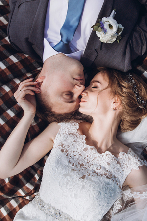 beautiful gorgeous bride and groom lying on tweed blanket in sunny spring park. happy wedding couple embracing and relaxing in green garden on picnic. happy marriage momentsの写真素材