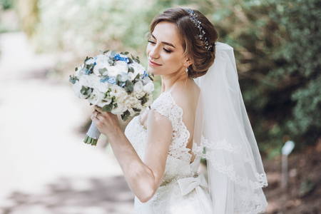 Beautiful blonde bride in elegant white wedding dress with white bouquet on a walk in the park after ceremonyの写真素材