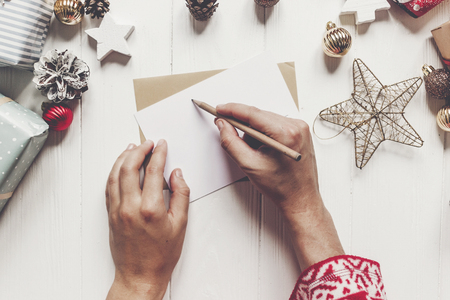 child writes letter to Santa Claus top view. kid hands holding pencil and making wish list and paper with christmas decorations and ornaments and presents on white wood. space for textの写真素材