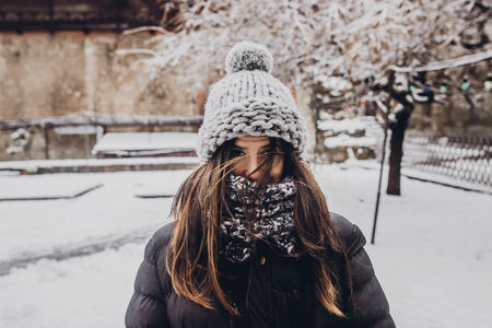 stylish hipster woman in knitted hat standing in snowy city street. beautiful fashionable girl in...