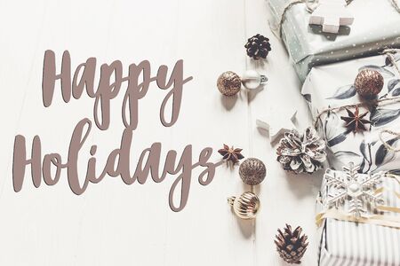 happy holidays text, seasonal greetings card sign. modern christmas wrapped presents with ornaments and toys on white wooden background top view,の写真素材