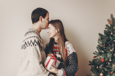 merry christmas and happy new year concept. stylish hipster couple in sweaters holding gift with red bow in room at christmas tree with lights and kissing. happy holidays. family  momentsの写真素材