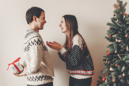 happy couple in stylish sweaters exchanging gifts in festive room at christmas tree. man holding surprise present behind back. merry christmas and happy new year conceptの写真素材