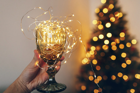 hand holding stylish vintage glass with light on background of beautiful christmas tree lights. merry christmas and happy new year concept. space for text. seasonal greetings, happy holidaysの写真素材