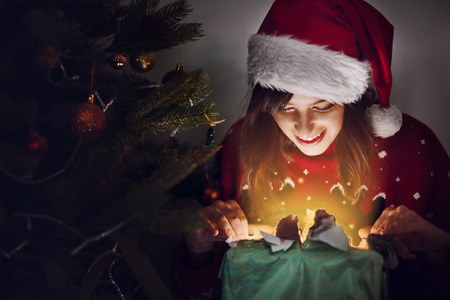 happy woman in santa hat opening gift box at christmas tree lights. magic light. surprise. space for text. merry christmas and happy new year concept. seasonal greetings, happy holidaysの写真素材